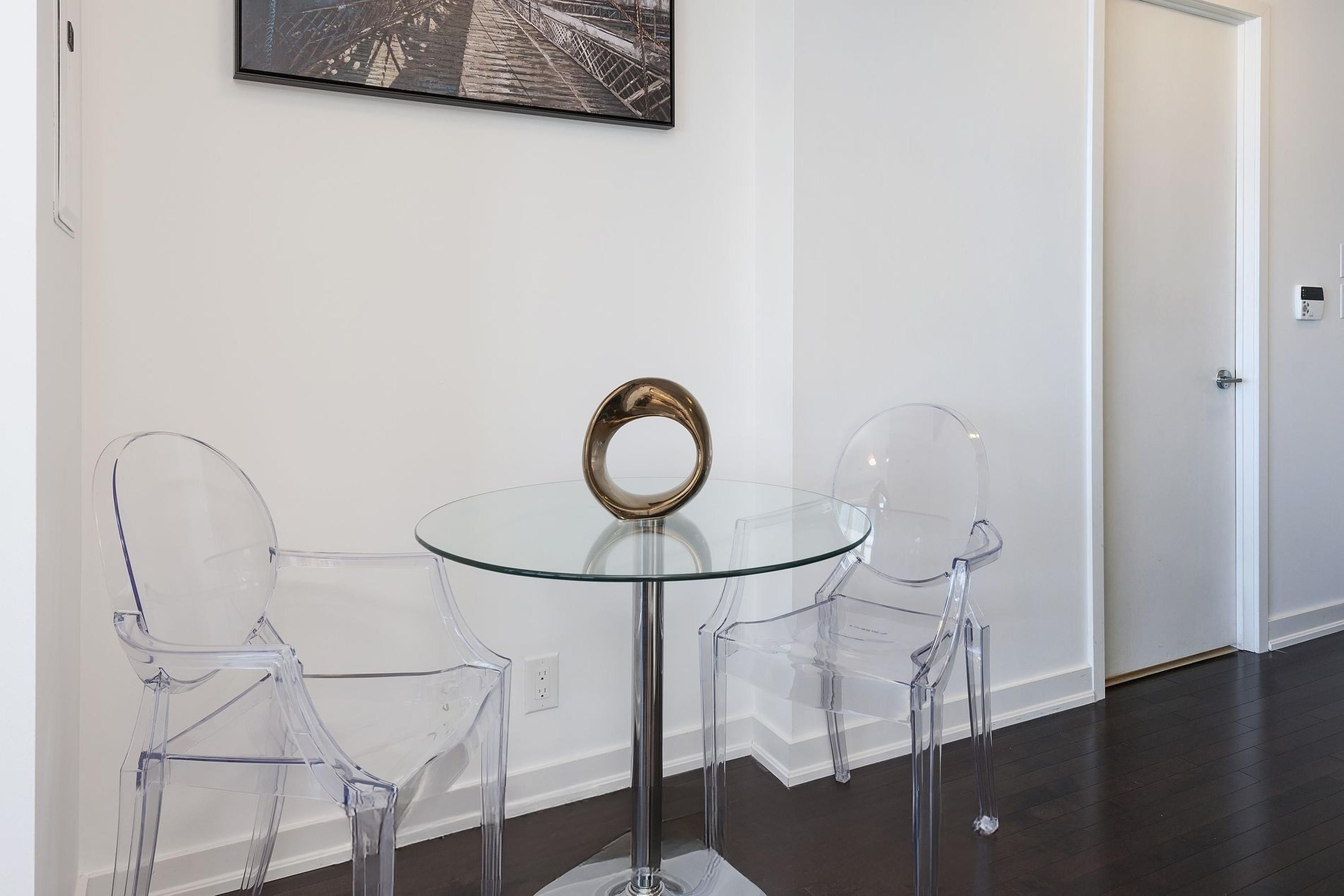 A modern room with a glass table and two clear chairs, a framed picture on the wall, and a door in the background.