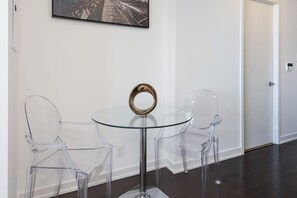 Quarto moderno com mesa de vidro e duas cadeiras transparentes, quadro na parede e porta ao fundo.