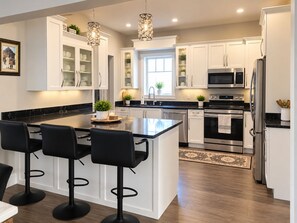 A modern kitchen with white cabinetry, stainless steel appliances, a central island with bar stools, pendant lighting, and a window providing natural light. -     Cottage Charm Meets Modern Comfort in the Heart of Woodstock (Woodstock)