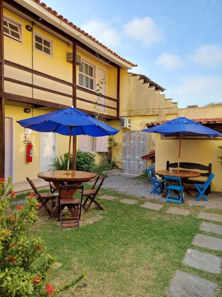 A courtyard with two wooden tables and blue umbrellas, surrounded by a yellow building with a tiled roof.