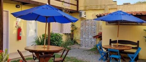 A courtyard with two wooden tables and blue umbrellas, surrounded by a yellow building with a tiled roof.