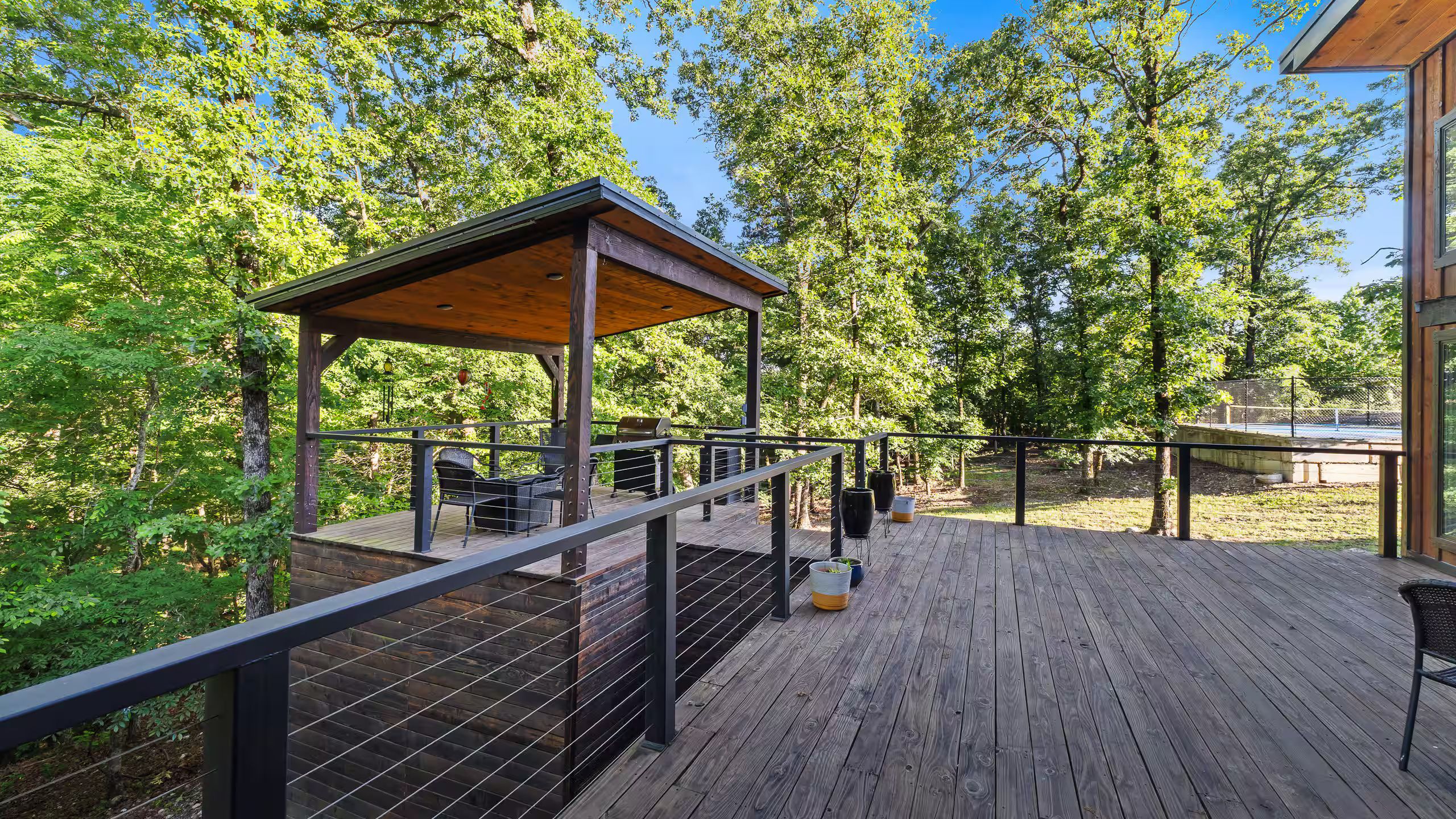 Une terrasse en bois dotée d’un espace salon couvert, de barbecues et offrant une vue sur une zone boisée.