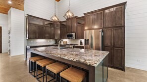 A modern kitchen with dark wood cabinets, a granite island with bar stools, stainless steel appliances, and pendant lighting. - Broken Bow Cabin w/ Hot Tub, Creek & 3 Acres (Broken Bow)