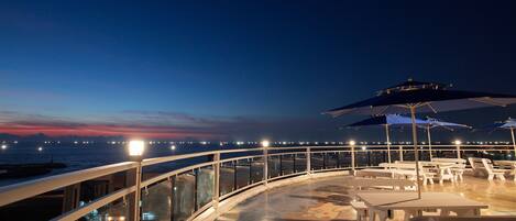 Una terraza con mesas y sillas, con vista a un cuerpo de agua y luces de la ciudad a lo lejos.