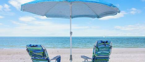 Deux chaises de plage sous un parasol bleu, sur une plage de sable, avec l’océan en arrière-plan.