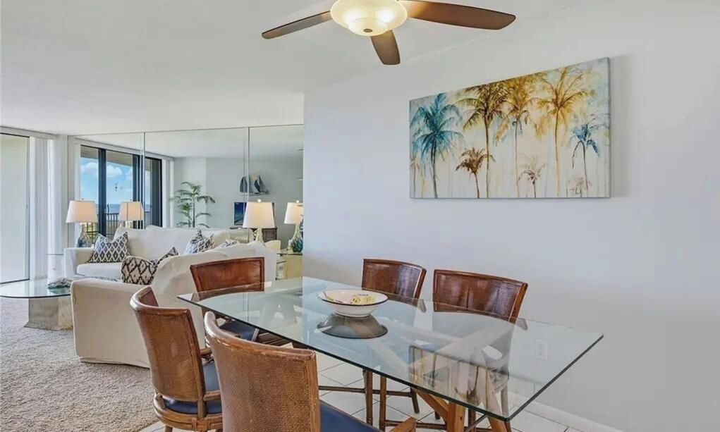 Un coin repas avec une table et des chaises en verre, un ventilateur de plafond et un tableau représentant des palmiers.