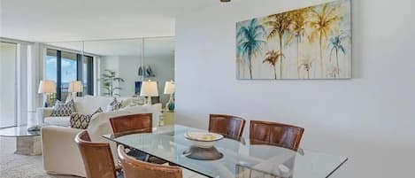 Un coin repas avec une table et des chaises en verre, un ventilateur de plafond et un tableau représentant des palmiers.