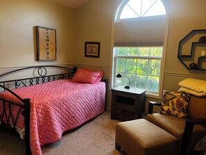 A bedroom with a bed, a desk, a chair, and a window with a view of greenery.