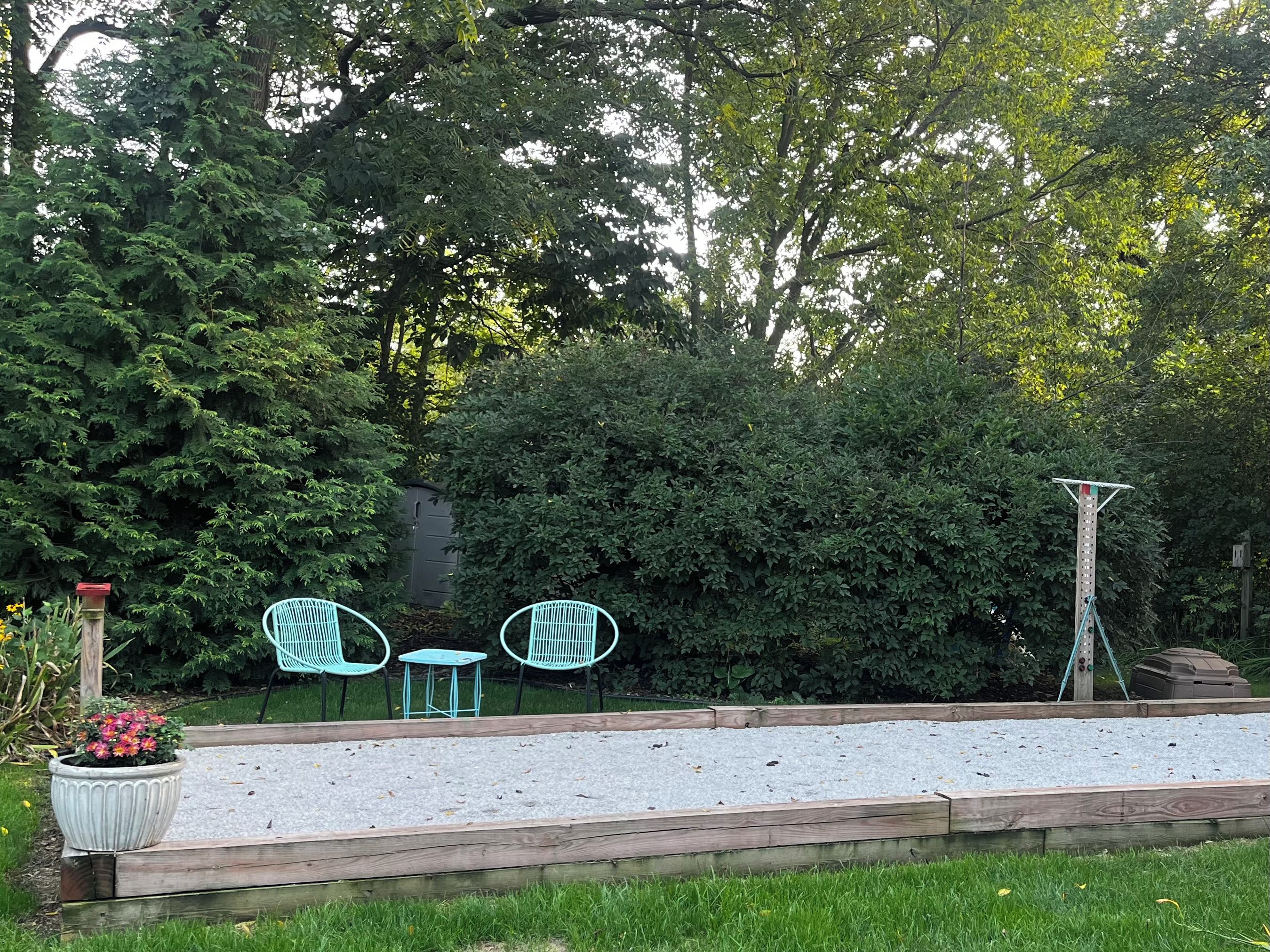 Un espace jardin agrémenté d’une terrasse en bois, de deux chaises vertes, d’une petite table et d’une structure haute.