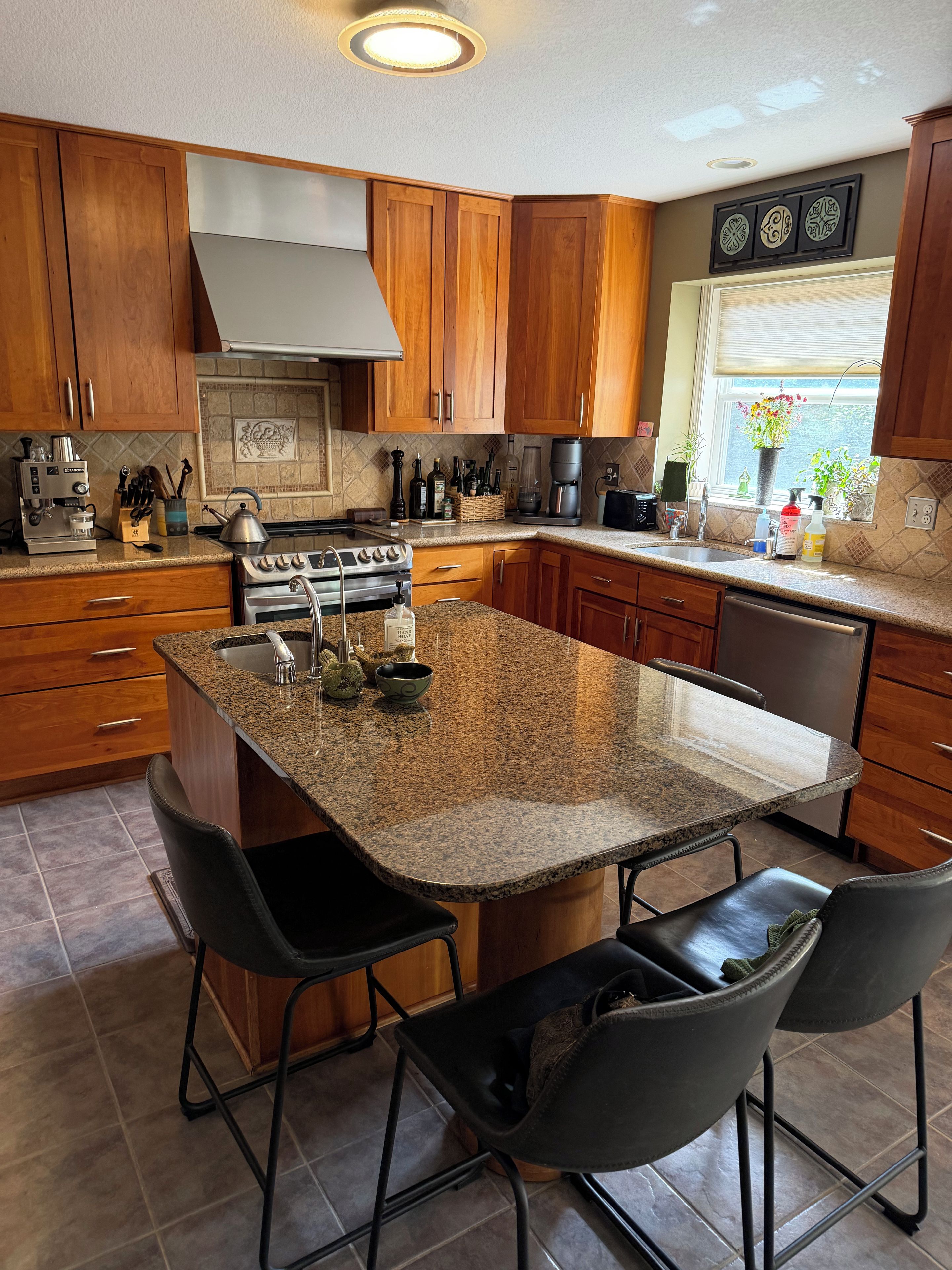 Een keuken met houten kasten, granieten aanrechtbladen, een centraal eiland met barstoelen en een raam met uitzicht.