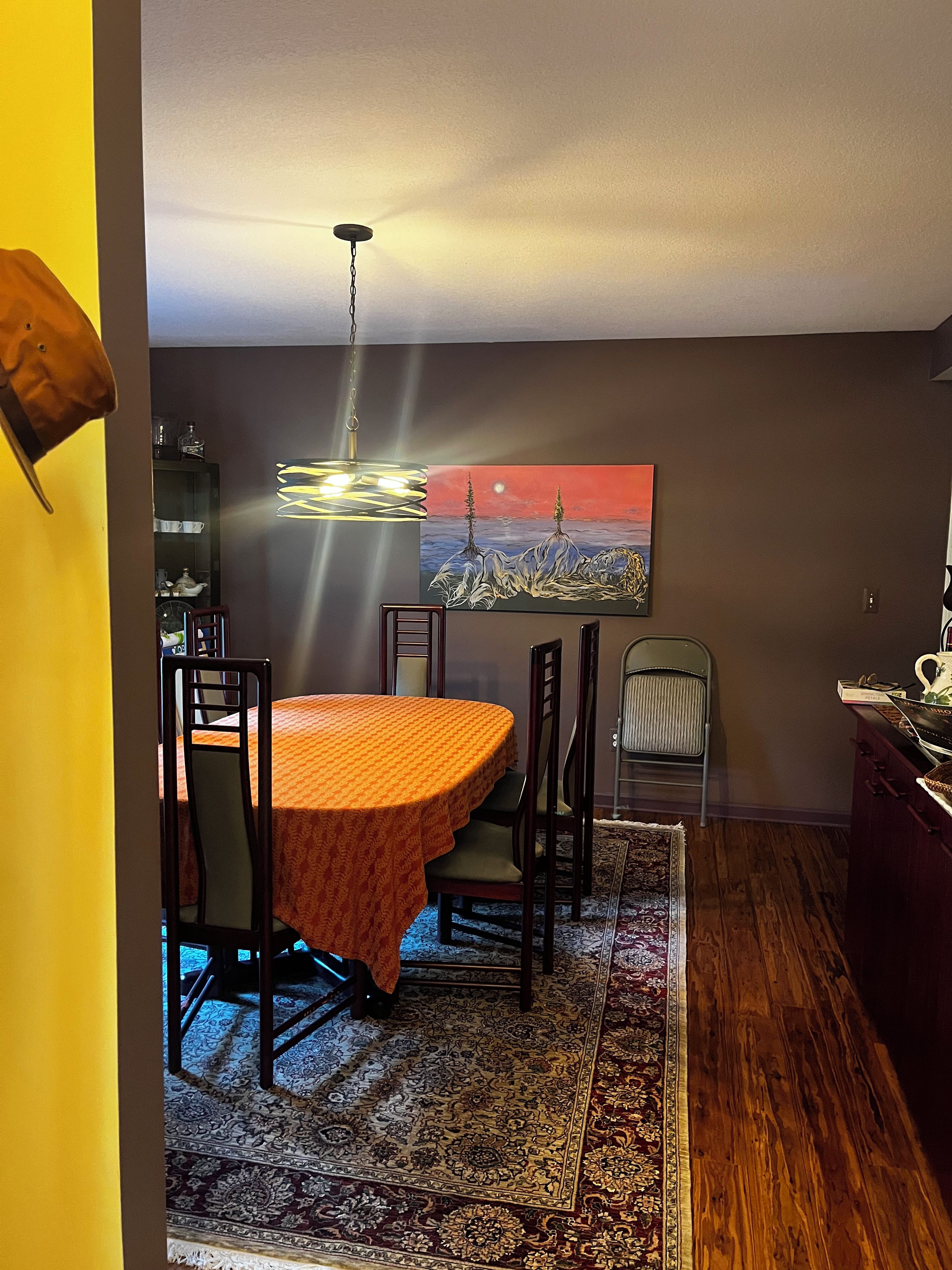 A dining area with a round table, chairs, a patterned rug, a painting on the wall, and a hanging light fixture.