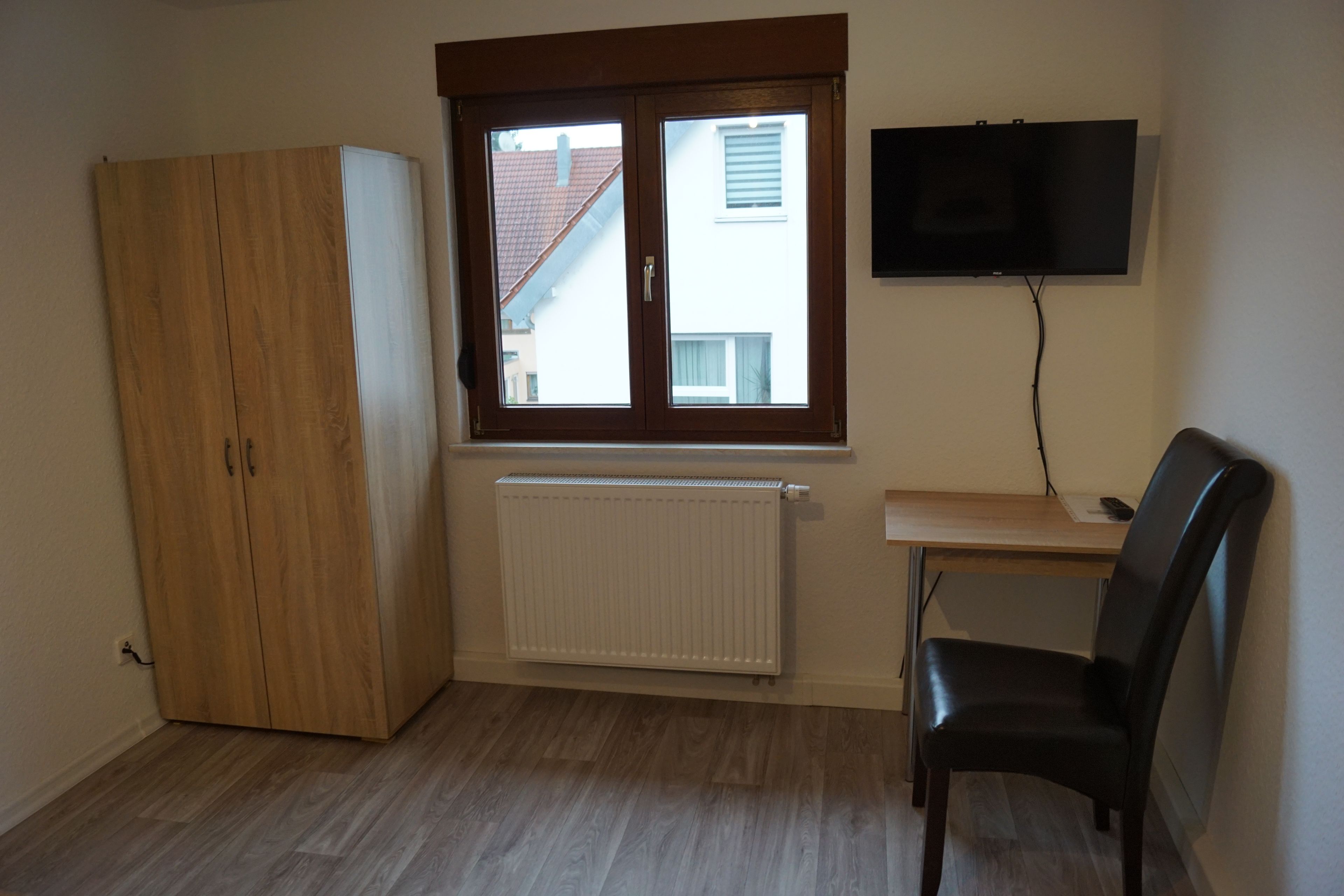 A room with a wooden wardrobe, a radiator, a desk with a chair, and a TV mounted on the wall.