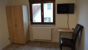 A room with a wooden wardrobe, a radiator, a desk with a chair, and a TV mounted on the wall.