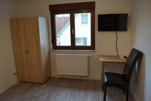 Quarto com guarda-roupa de madeira, radiador, escrivaninha com cadeira e televisão fixada na parede.