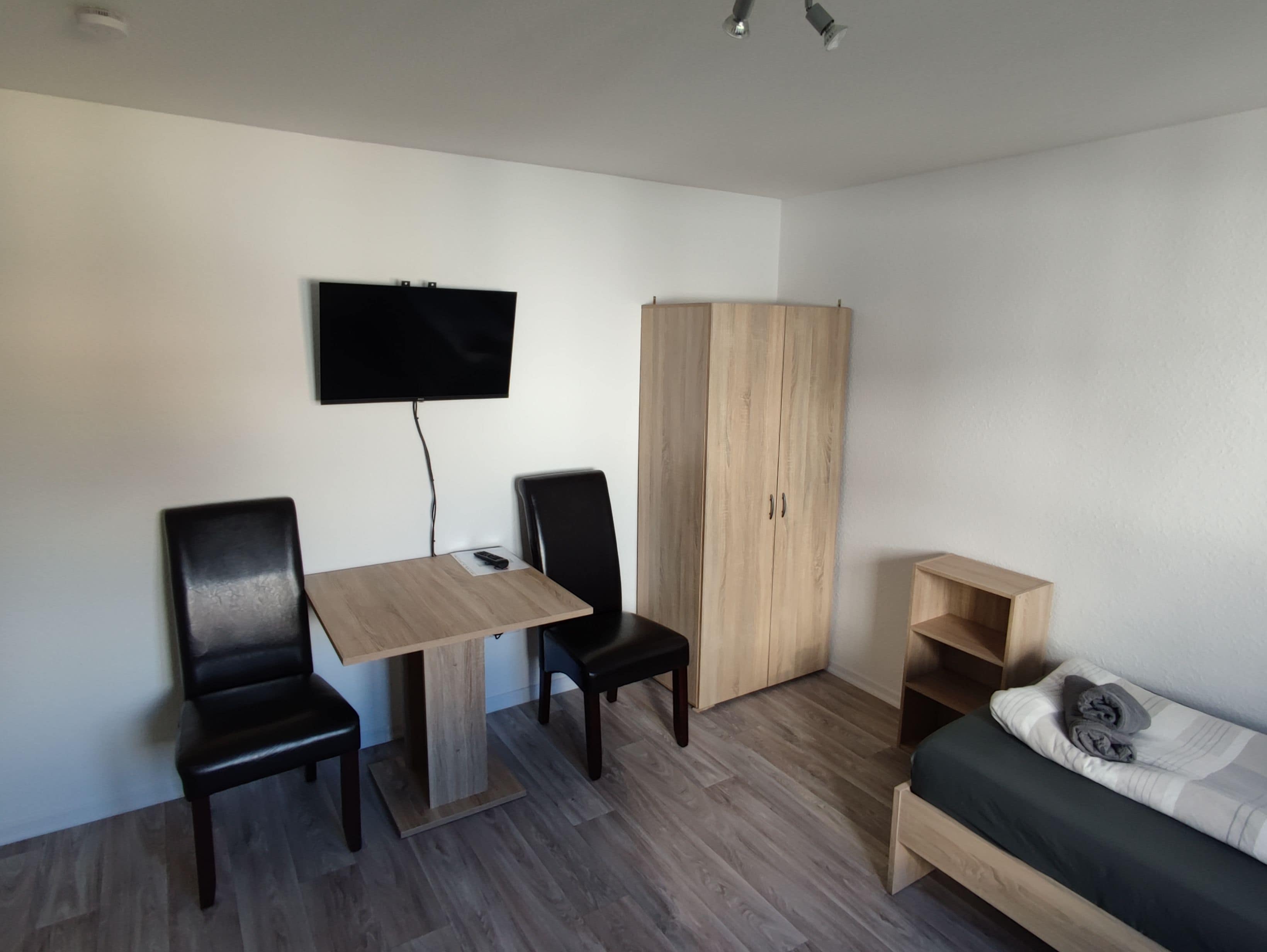 A small, modern room with a wooden table, two chairs, a television mounted on the wall, a wardrobe, and a bed with a pillow.