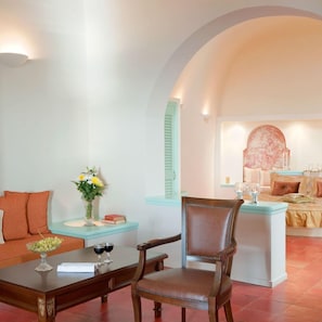 A living room with a sofa, a coffee table, a chair, a small table with a vase of flowers, and a bed with a decorative headboard. - Vallas Apartments (Santorini)