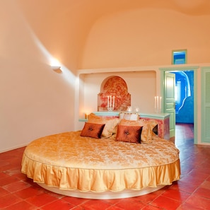 A circular bed with a ruffled edge, a bedside table with candles, and a view of a blue room through an open door. - Vallas Apartments (Santorini)