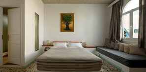 Habitación de hotel con cama, lámparas de noche, ventana con cortinas, un cuadro de un árbol y un banco con cojines.