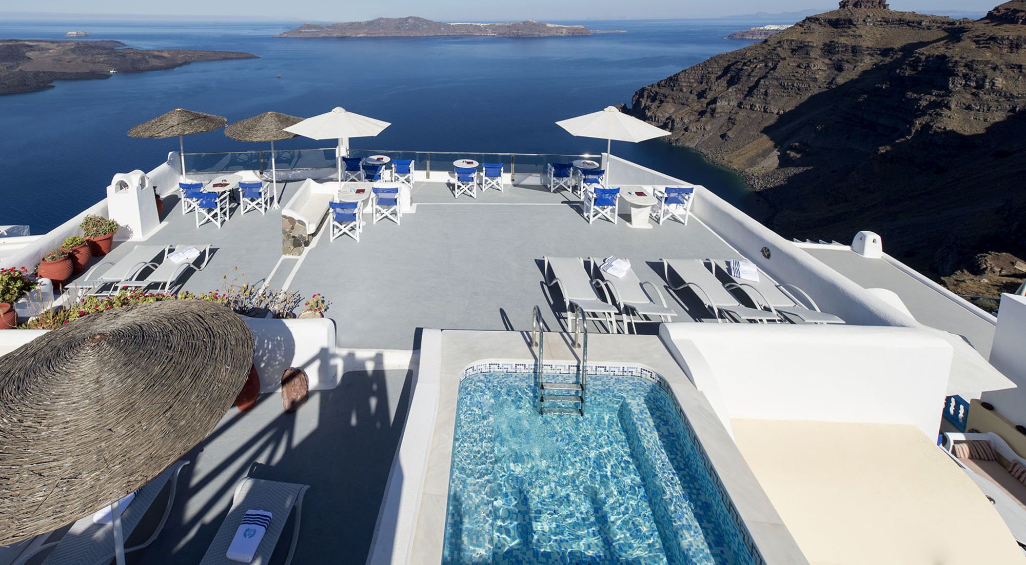 A rooftop pool area with lounge chairs and umbrellas, overlooking a body of water and rocky terrain.