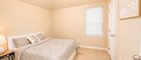 Un dormitorio con cama, mesita de noche, ventana con persianas y puerta.
