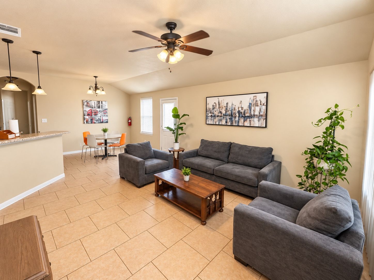 A spacious living area with a gray sofa, a coffee table, and a ceiling fan. There is a dining area with a table and chairs, and a kitchen area with a counter and appliances.
