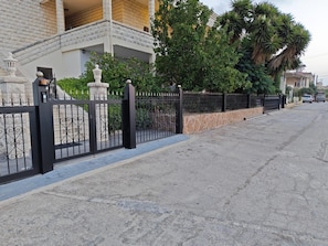 Un área residencial cerrada con camino pavimentado, cerca metálica y un edificio con balcón.