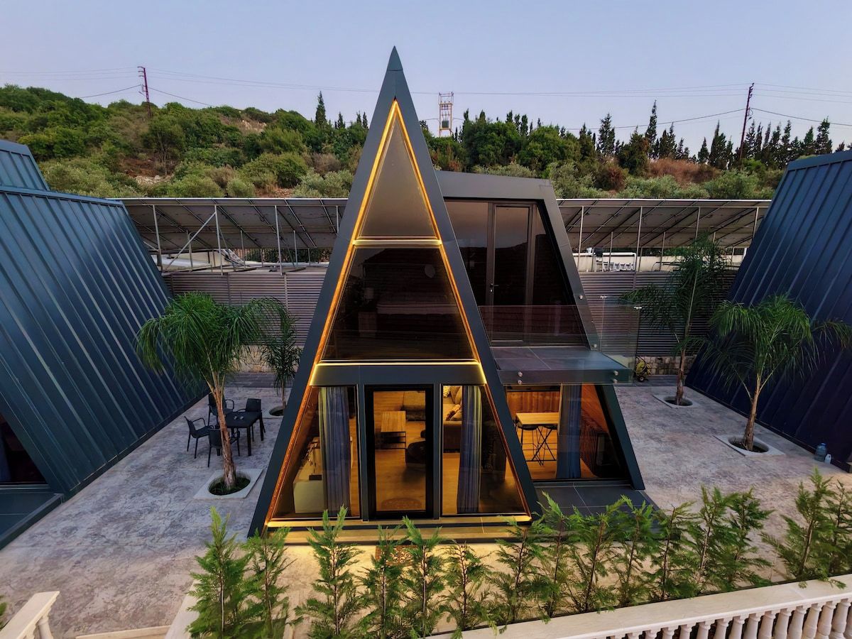 Un edificio moderno de forma triangular con fachada de vidrio, rodeado de vegetación y un área exterior cubierta con asientos.