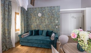 Una habitación con un sofá color turquesa, suelo de madera, papel tapiz estampado y un jarrón con flores.