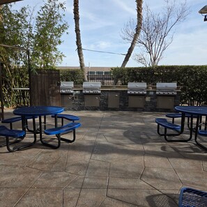 An outdoor patio area with barbecue grills, blue picnic tables, and a stone-tiled floor.