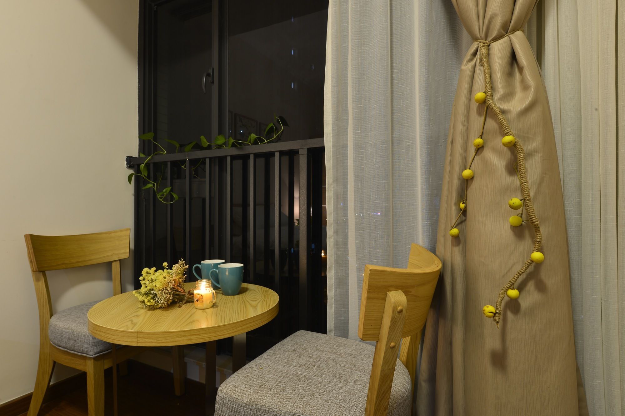 Een klein balkon met twee houten stoelen, een ronde tafel met een kaars en een blauwe mok, en een gordijn met gele kralen.