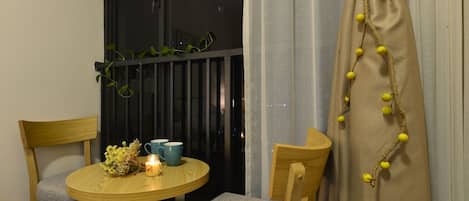 Een klein balkon met twee houten stoelen, een ronde tafel met een kaars en een blauwe mok, en een gordijn met gele kralen.