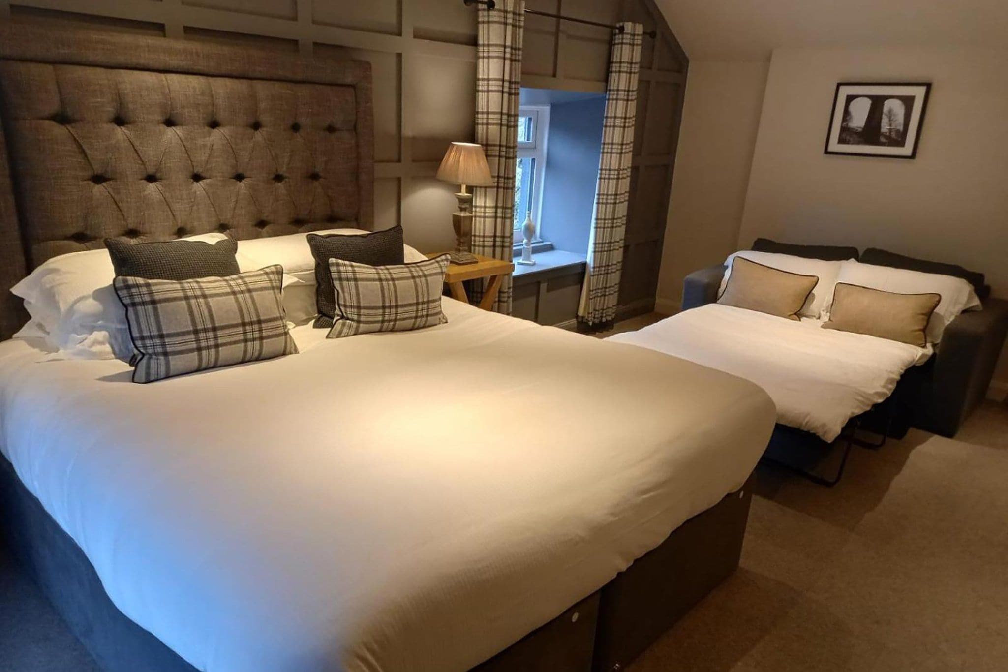 A bedroom with a large bed, a smaller bed, a nightstand, a lamp, and a window with curtains.