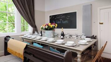 A dining area with a long table set for a meal, a chalkboard with a welcome message, and a window with curtains.