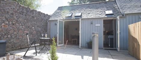 A stone wall, a patio with a table and chairs, a small potted plant, and a building with an open door.