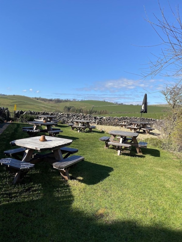 An outdoor setting with multiple picnic tables arranged on a grassy field, surrounded by stone walls and a clear sky.
