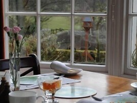 A dining table with a vase of flowers, a glass of orange juice, and a bird feeder outside the window.