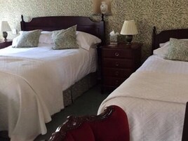 A hotel room with two beds, each with a wooden headboard and white bedding. There is a nightstand with a lamp between the beds.