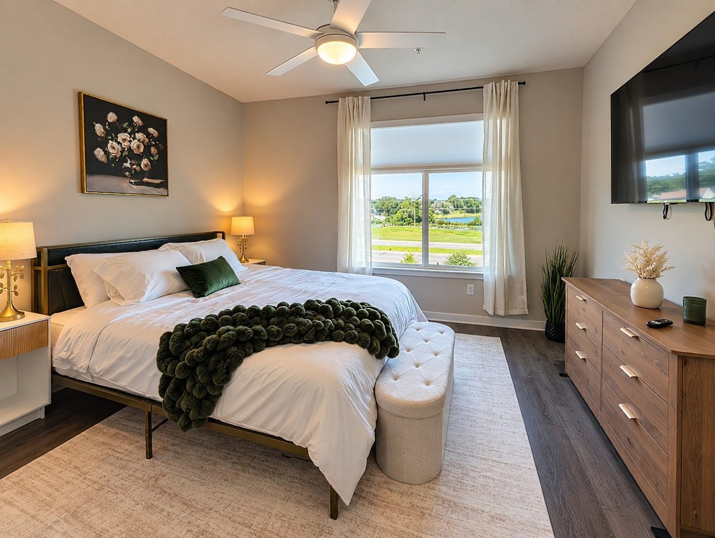 A bedroom with a bed, a ceiling fan, a window with curtains, a television, and a bedside table with a lamp.