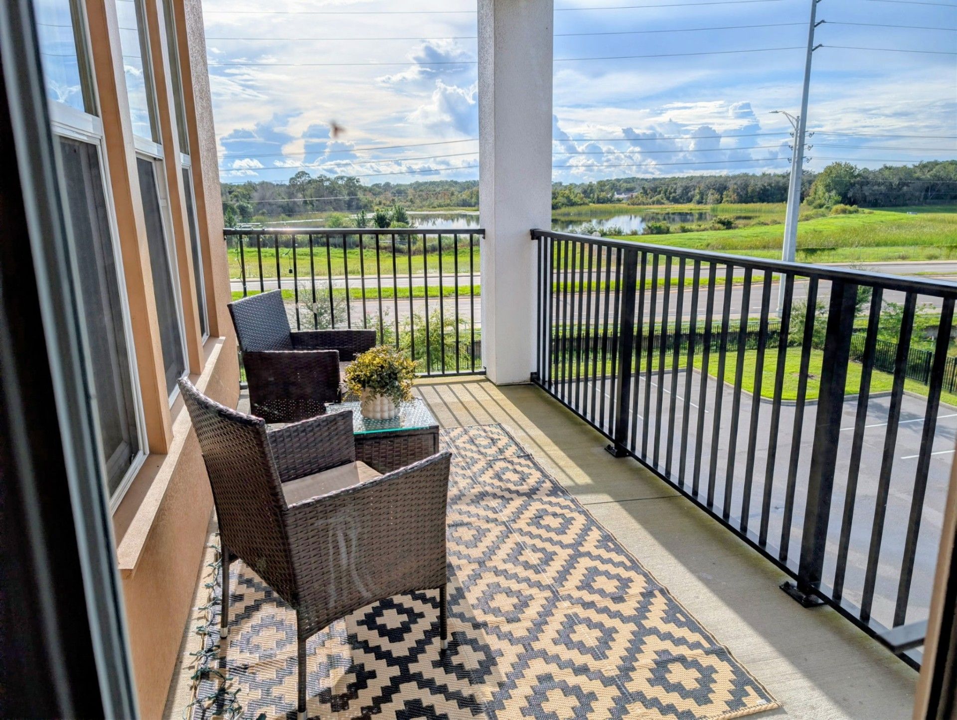 A balcony with wicker furniture and a patterned rug, overlooking a green landscape and a body of water.