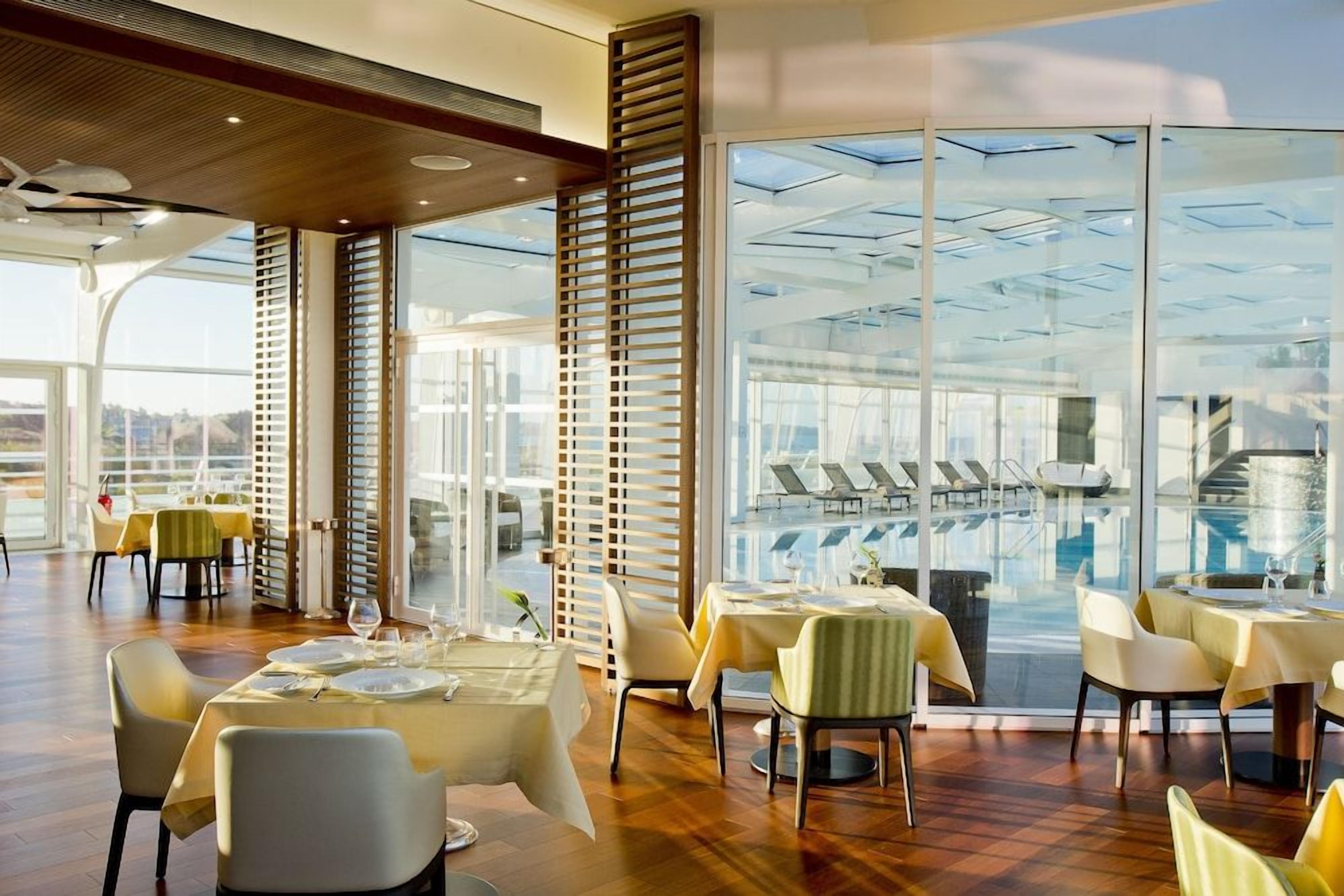 Un restaurant avec des tables dressées pour le service, de grandes fenêtres et une vue sur un espace piscine.