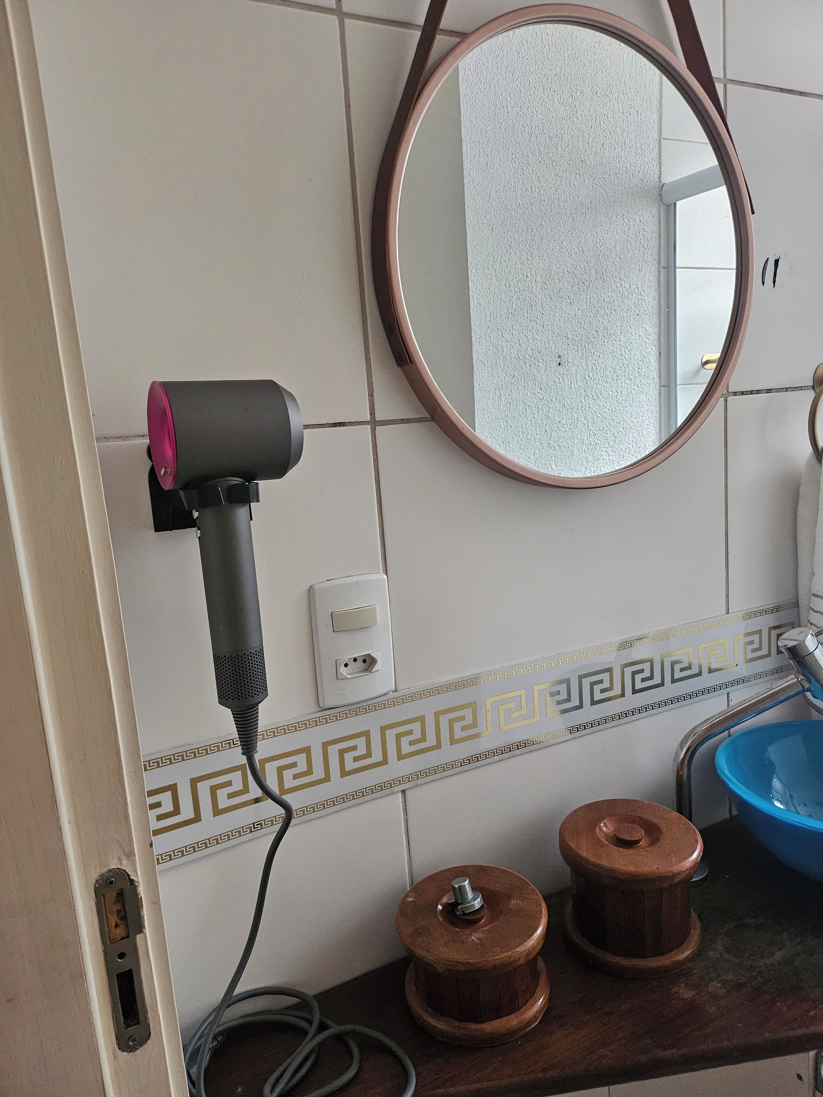 Casa de banho com secador de cabelo fixado na parede, espelho redondo, lavatório com cuba azul e azulejos decorativos.