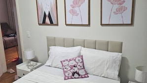 Una cama bien hecha con edredón blanco y manta rosa, una mesita de noche con l ámpara, y una pared con tres cuadros enmarcados.