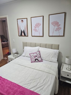 A neatly made bed with a white comforter and a pink blanket, a bedside table with a lamp, and a wall with three framed artworks. - ROSE DE OLIVEIRA CESARI (Teresópolis)