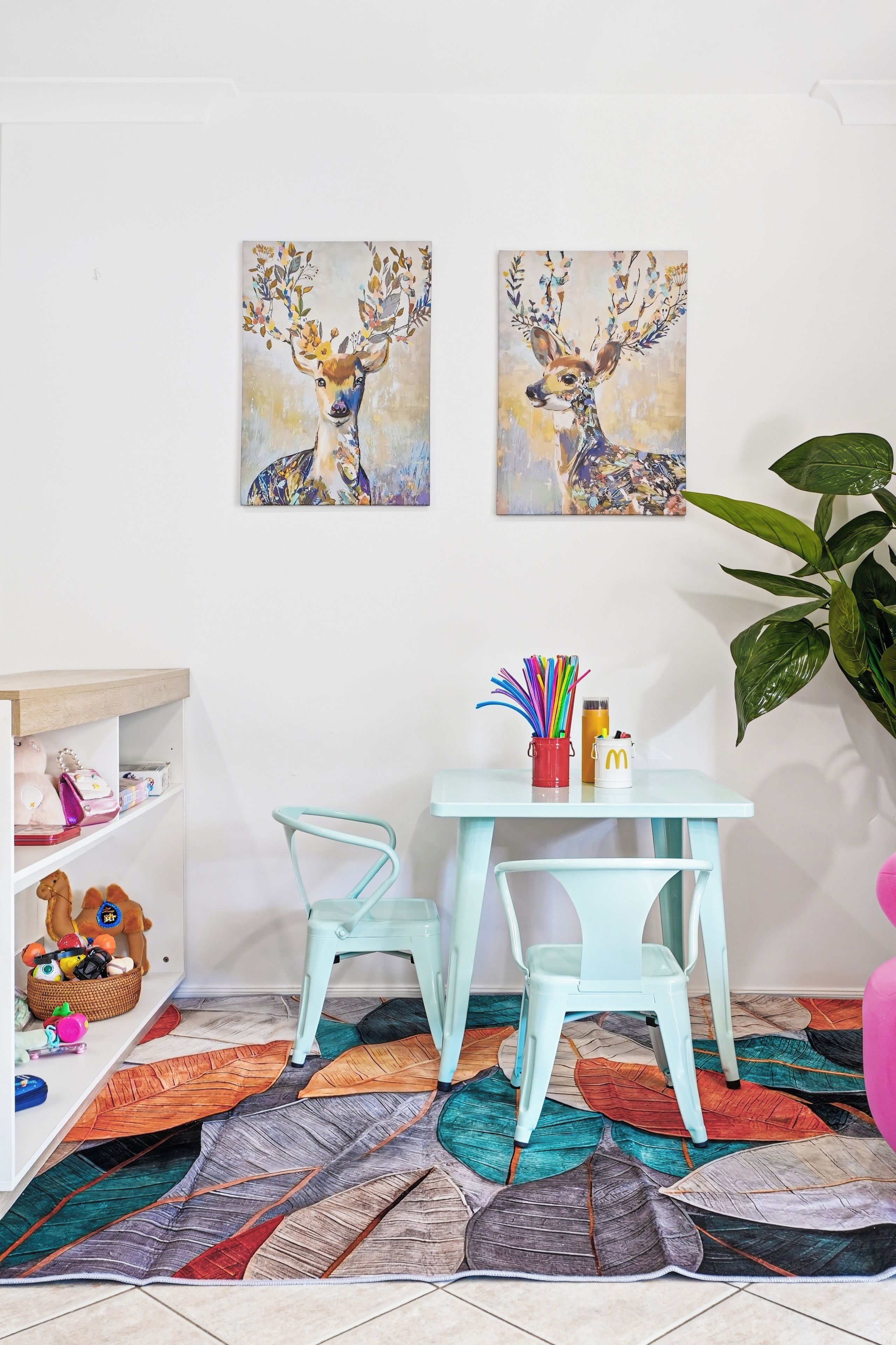 Una habitación con una mesa color turquesa, dos sillas, un tapete con estampado de hojas coloridas, un estante con peluches y dos cuadros de ciervos colgados en la pared.