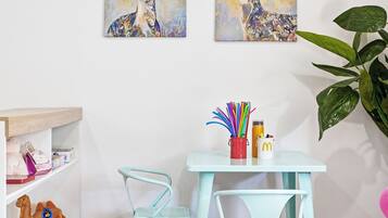 Una habitación con una mesa color turquesa, dos sillas, un tapete con estampado de hojas coloridas, un estante con peluches y dos cuadros de ciervos colgados en la pared.