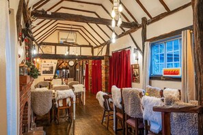 A rustic interior with exposed wooden beams, a brick fireplace, and a dining area with chairs covered in fur. - The Bull at The Green (Lewes)