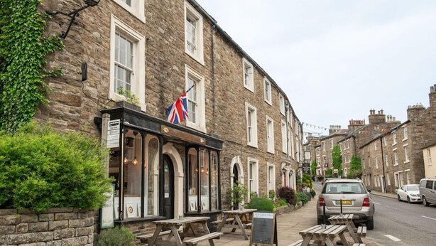 A stone building with a flag, outdoor seating, and parked cars. - Milton House Bed and Breakfast (Leyburn)