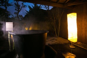 Una olla grande y un aparato de cocina vertical, ambos despidiendo vapor, en un entorno al aire libre con árboles y una estructura de madera al fondo.
