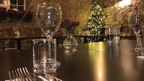A dining table set with cutlery, a glass, and a vase with flowers, with a Christmas tree in the background.