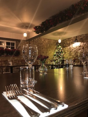 A dining table set with cutlery and wine glasses, a Christmas tree in the background, and a stone wall. - The George At Nunney (Frome)